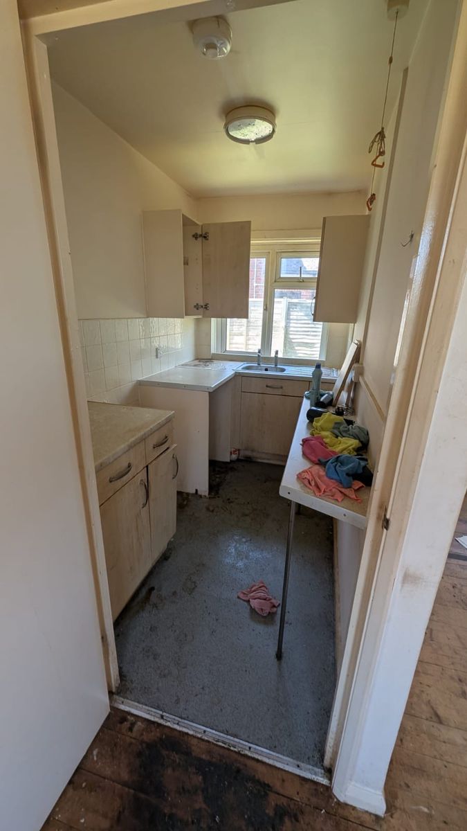 A narrow kitchen seen through a doorway, with cupboards, a sink under the window and cloths on a side shelf