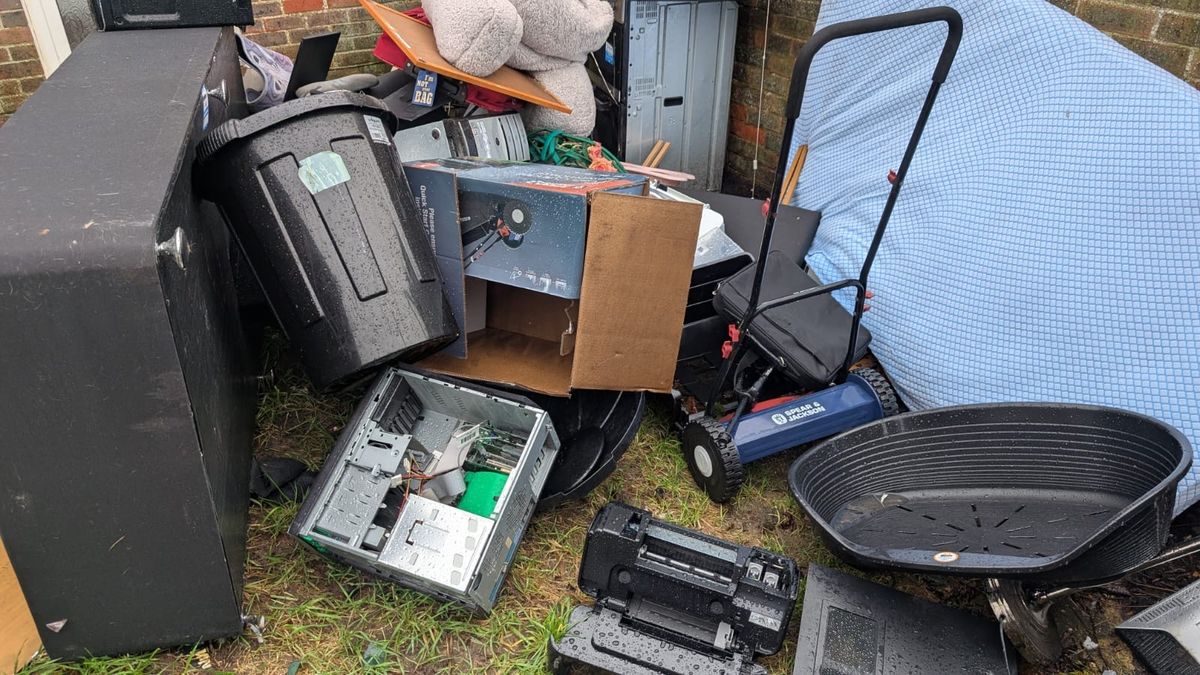Bulky household items gathered on the grass outside a house: a mattress, wheelie bin, computer tower, TV, lawnmower and plastic pet bed
