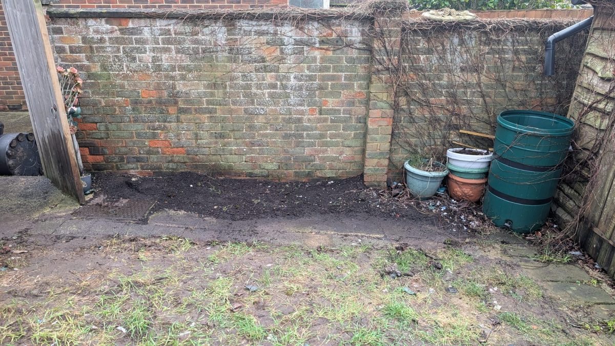A back garden cleared to bare soil beside a brick wall with a few plant pots left