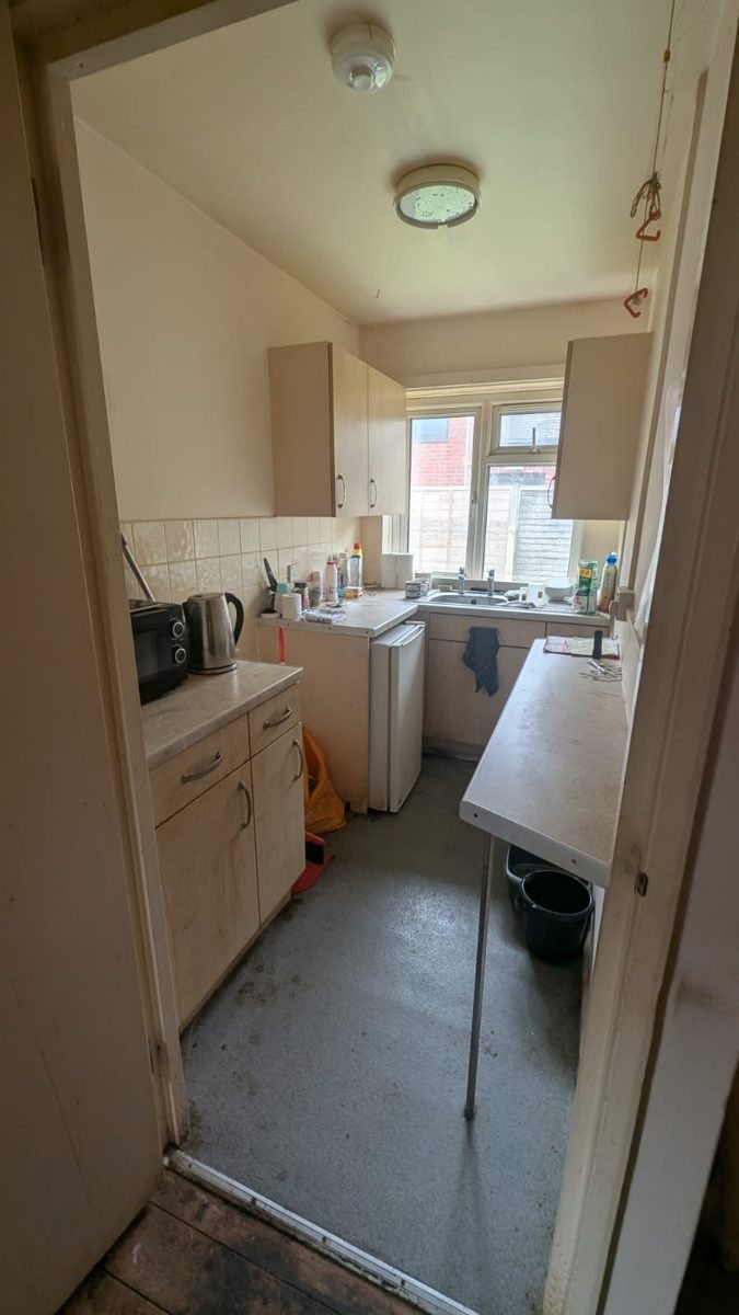 A small kitchen seen through a doorway with a microwave and clutter on the worktop, before clearance