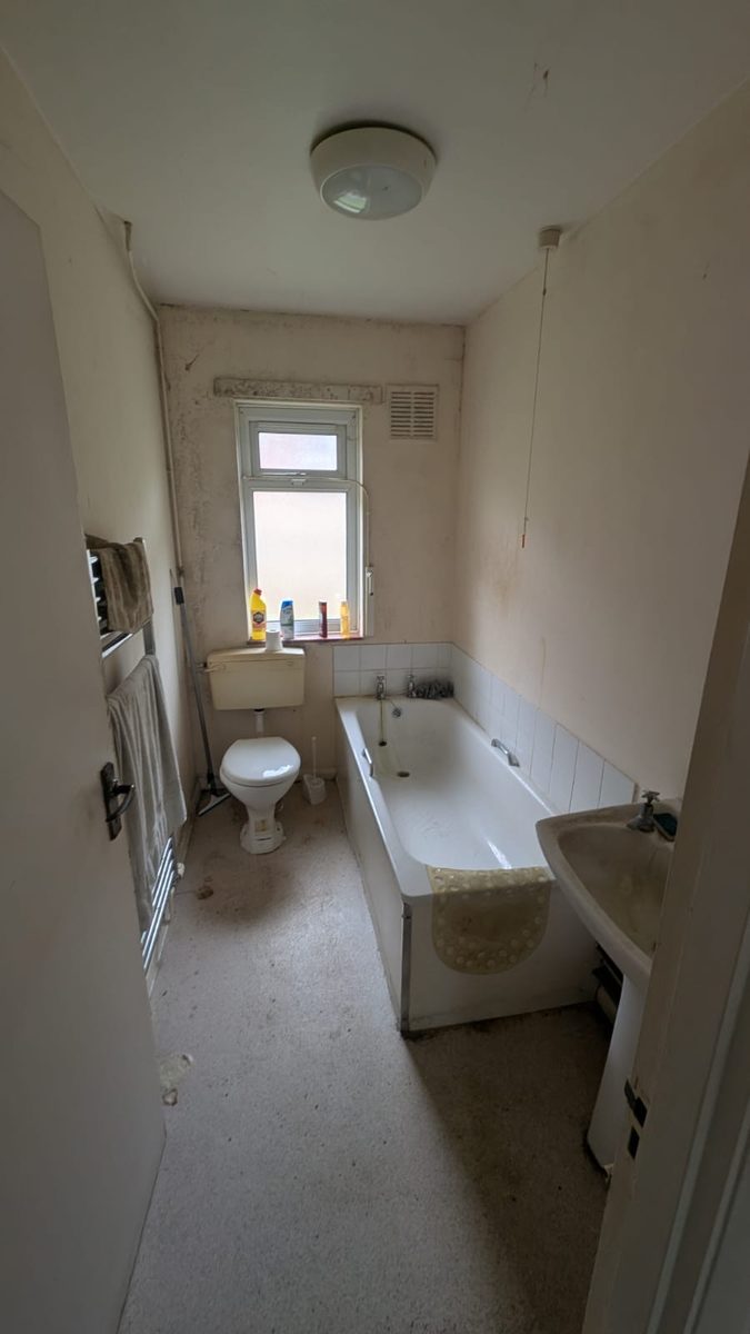A small bathroom seen from the doorway with towels, a bath mat, bottles on the windowsill and a brush beside the toilet