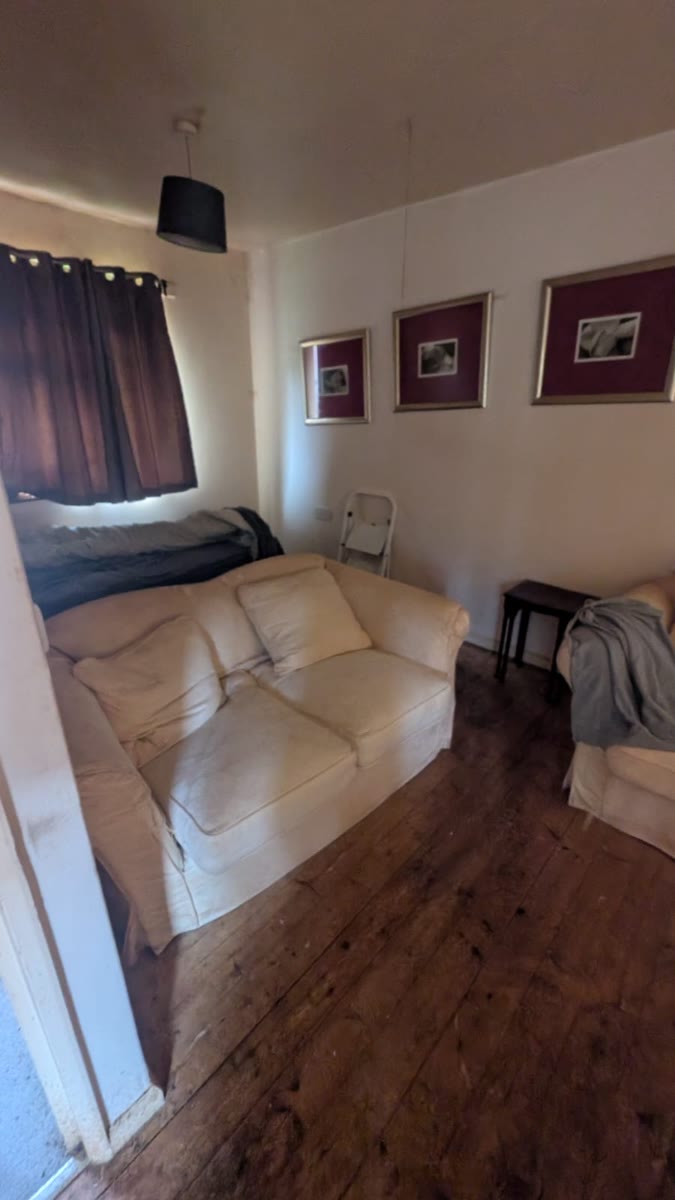 A room with a bed and mattress under the window, a cream two-seater sofa, an armchair and framed pictures on the wall