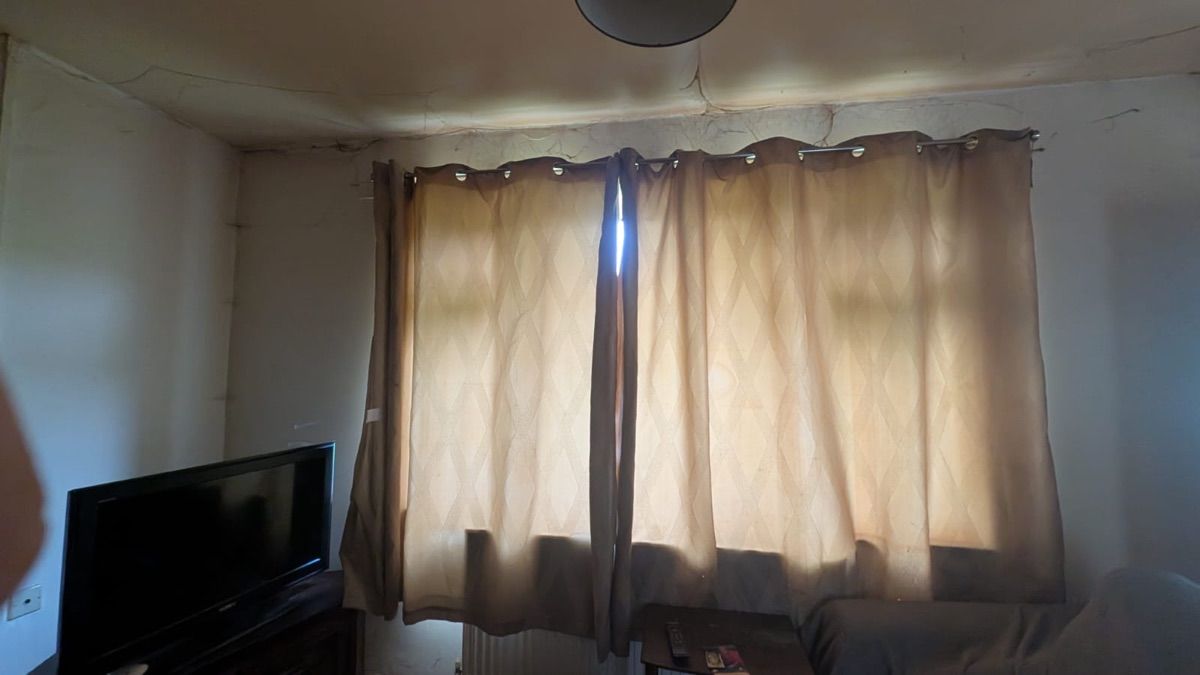 A living room before clearance, with a grey fabric sofa, a television on a low unit and patterned curtains drawn across the window